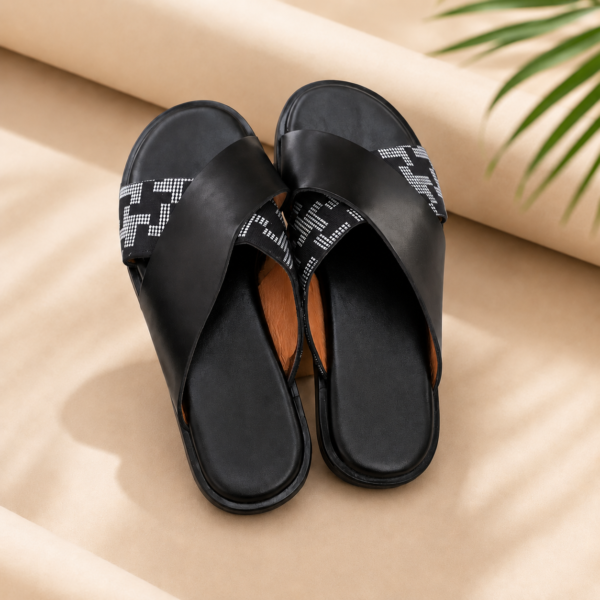 Mules croisées unisexes noires avec lanières graphiques blanches et noires sur fond beige.
